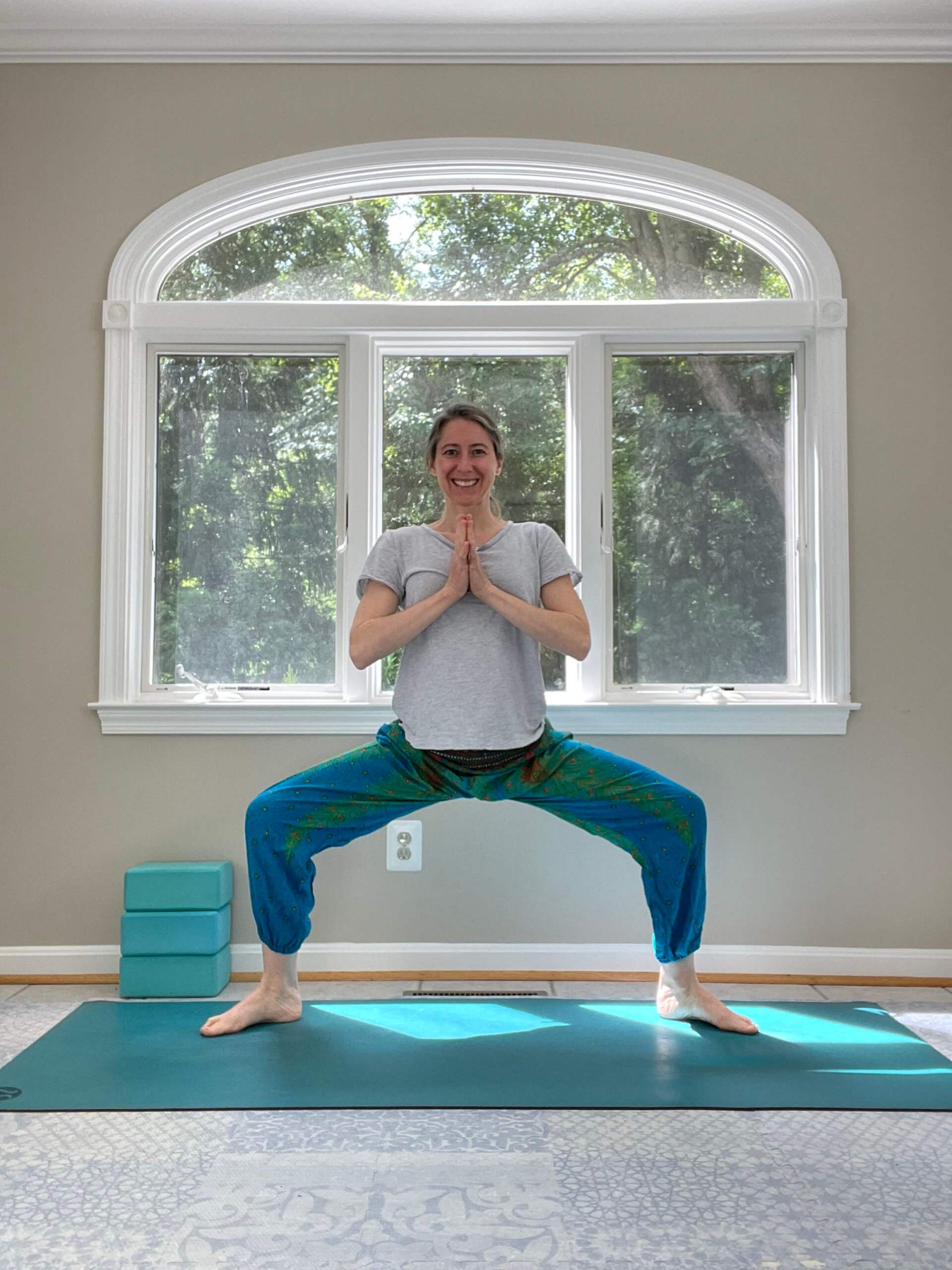 A woman in blue flowy pants and a blue top in Goddess Pose on a turquoise yoga mat